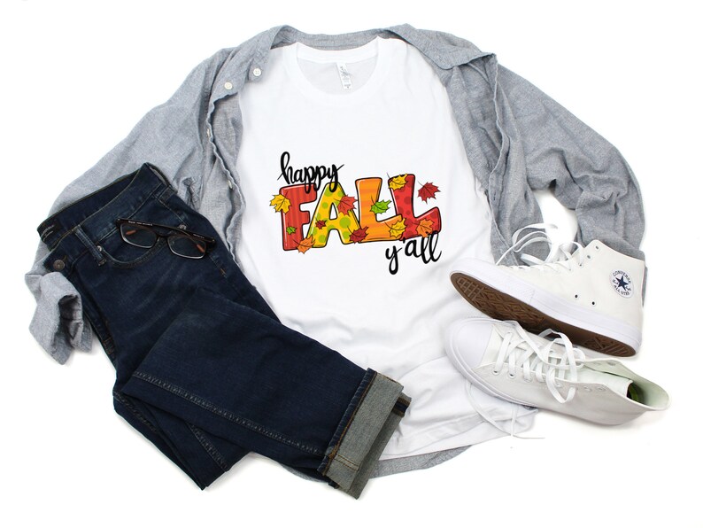 May include: A white t-shirt with the words "happy FALL y'all" in colorful letters and fall leaf accents. The shirt is paired with blue jeans, a gray button-down shirt, and white sneakers.