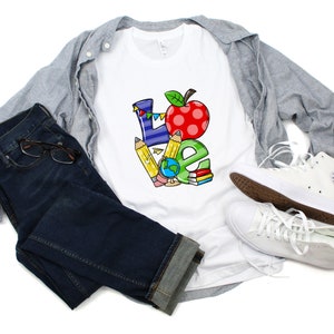 May include: White t-shirt with a colorful "LOVE" graphic. The design incorporates a red apple, pencils, a globe, and books. The shirt is paired with blue jeans, a gray button-down shirt, and white sneakers.