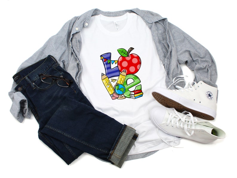 May include: White t-shirt with a colorful "LOVE" graphic. The design incorporates a red apple, pencils, a globe, and books. The shirt is paired with blue jeans, a gray button-down shirt, and white sneakers.