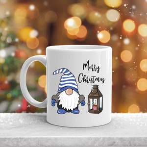 May include: White ceramic mug featuring a cartoon gnome with a blue and white striped hat, blue gloves, and a white beard. The mug also displays the words "Merry Christmas" and a lantern illustration. Festive holiday theme.