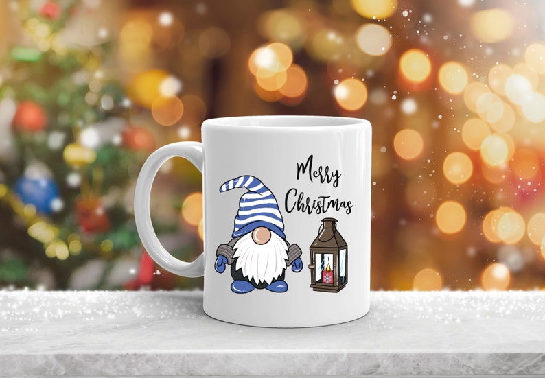 May include: White ceramic mug featuring a cartoon gnome with a blue and white striped hat, blue gloves, and a white beard. The mug also displays the words "Merry Christmas" and a lantern illustration. Festive holiday theme.