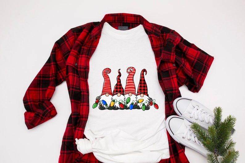 以下が含まれることがあります： 赤いチェック柄のシャツの上に置かれた白いTシャツ。Tシャツには、赤と黒の帽子をかぶった3人のノームがクリスマスの電飾に囲まれているデザインがプリントされています。