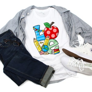 May include: White t-shirt with a colorful graphic of the word "LOVE" made up of school supplies like pencils, a globe, and a red apple. The shirt is styled with blue jeans, a gray button-down shirt, and white sneakers.