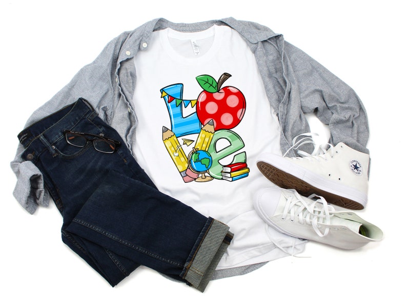 May include: White t-shirt with a colorful graphic of the word "LOVE" made up of school supplies like pencils, a globe, and a red apple. The shirt is styled with blue jeans, a gray button-down shirt, and white sneakers.