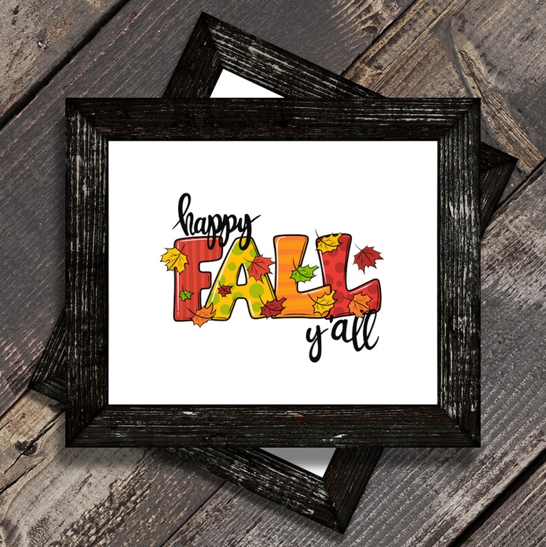 May include: A framed print with the words "happy FALL y'all" in colorful, stylized letters. The letters are decorated with fall leaves. The frame is black and has a distressed wood appearance. The background is a wooden surface.