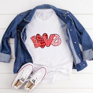 以下が含まれることがあります： 赤とピンクのデザインで「LOVE」という文字が入った白いTシャツ、デニムジャケットと白いスニーカーを組み合わせたもの。「LOVE」という文字は、水玉模様とストライプが入ったバブルレタースタイルです。