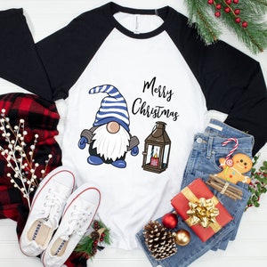 May include: White and black raglan-style shirt with a Christmas gnome graphic. The gnome wears a blue and white striped hat and has a white beard. The words "Merry Christmas" are written above a lantern.