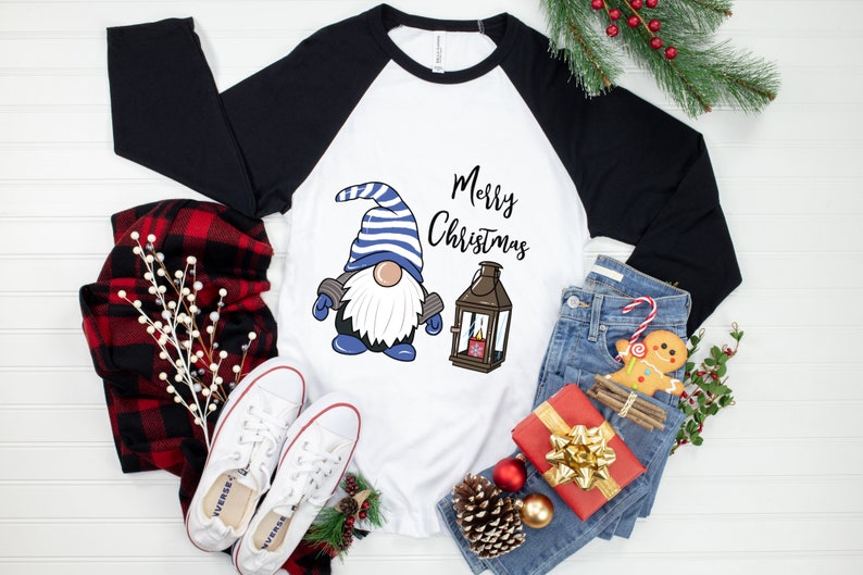 May include: White and black raglan-style shirt with a Christmas gnome graphic. The gnome wears a blue and white striped hat and has a white beard. The words "Merry Christmas" are written above a lantern.