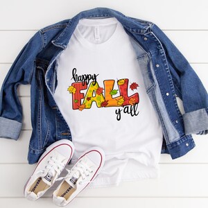 May include: White t-shirt with the text "happy FALL y'all" in colorful letters and autumn leaves. The shirt is paired with a blue denim jacket and white canvas sneakers with red accents, creating a casual fall ensemble.