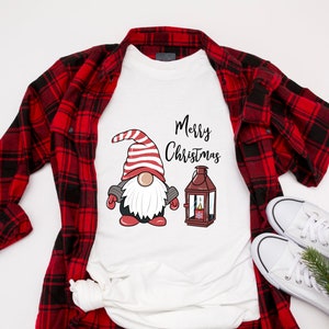May include: White t-shirt with a Christmas gnome graphic and the words "Merry Christmas". The gnome wears a red and white striped hat and mittens. A red and black plaid shirt and white sneakers are also in the image.