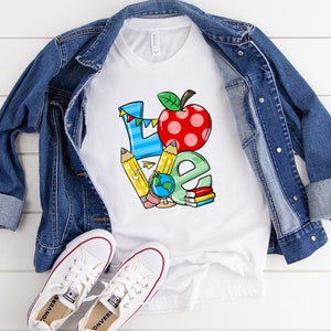 May include: White t-shirt with a colorful graphic of the word "LOVE" with a red apple and school supplies. The shirt is laid on a white wooden surface with a denim jacket and white sneakers.