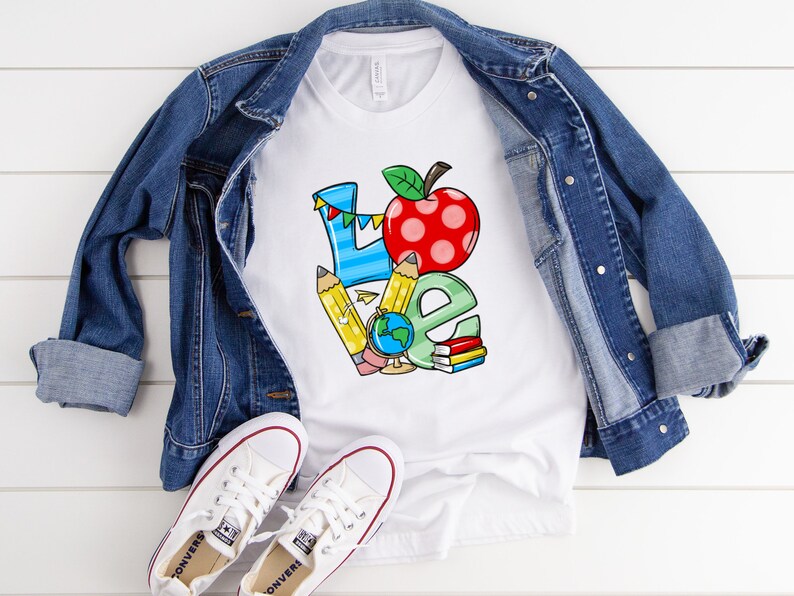 May include: White t-shirt with a colorful graphic of the word "LOVE" with a red apple and school supplies. The shirt is laid on a white wooden surface with a denim jacket and white sneakers.