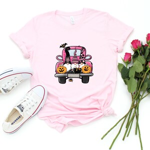 May include: A pink t-shirt with a graphic of three gnomes riding in a pink truck with pumpkins and bats. The gnomes are wearing hats and have beards. The truck has a black outline and black wheels.