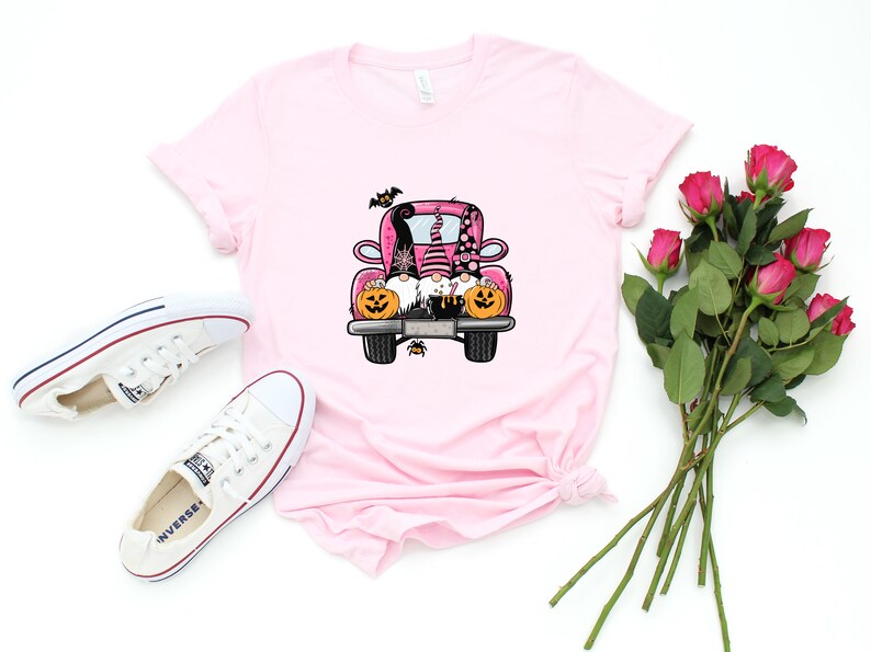 May include: A pink t-shirt with a graphic of three gnomes riding in a pink truck with pumpkins and bats. The gnomes are wearing hats and have beards. The truck has a black outline and black wheels.