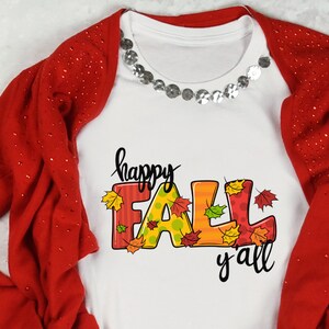May include: White t-shirt with the words "happy FALL y'all" in black script and colorful block letters. The letters are decorated with fall leaves. A red cardigan with sparkly details is draped over the shirt.