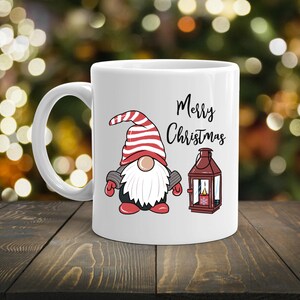 May include: White ceramic mug featuring a Christmas gnome with a red and white striped hat, a white beard, and the words "Merry Christmas." A red lantern is also depicted. The mug is on a wooden surface.