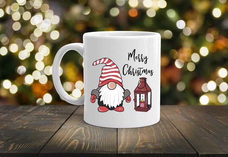 May include: White ceramic mug featuring a Christmas gnome with a red and white striped hat, a white beard, and the words "Merry Christmas." A red lantern is also depicted. The mug is on a wooden surface.