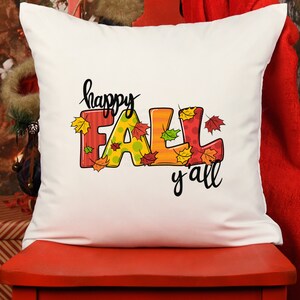 May include: A white square pillow with the words "happy FALL y'all" in black script. The word "FALL" is in colorful letters with fall leaves. The pillow is on a red surface.