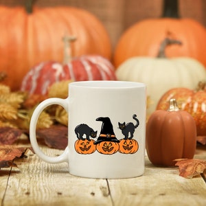 May include: White ceramic mug featuring a Halloween design. The design includes three orange jack-o'-lanterns, two black cats, and a witch's hat. The background includes pumpkins and autumn leaves, creating a festive fall scene.