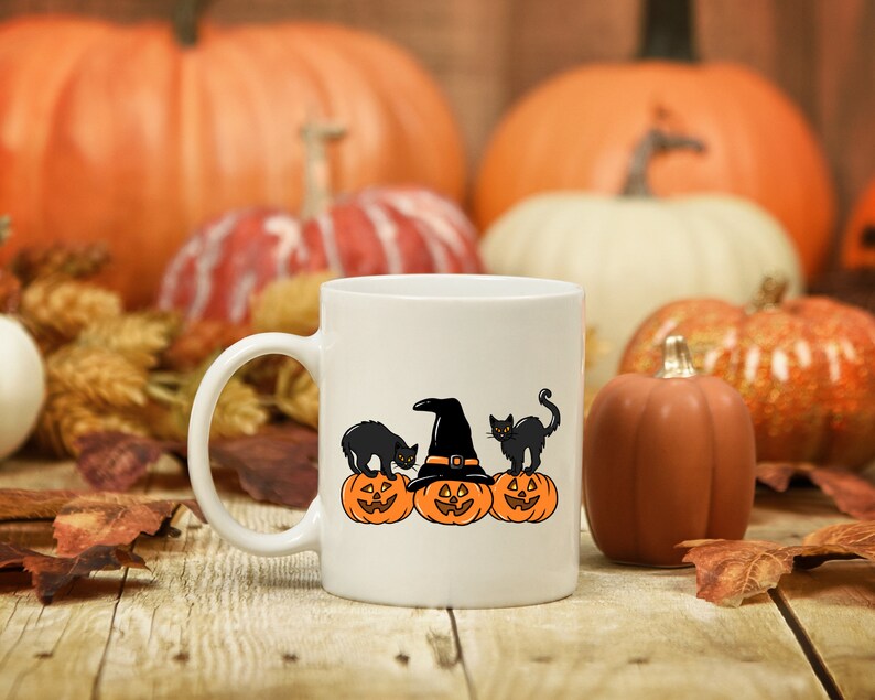 May include: White ceramic mug featuring a Halloween design. The design includes three orange jack-o'-lanterns, two black cats, and a witch's hat. The background includes pumpkins and autumn leaves, creating a festive fall scene.