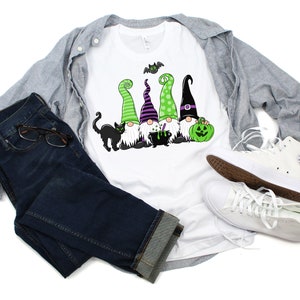 May include: White t-shirt featuring a Halloween gnome design with green, purple, and black hats, a black cat, and a green pumpkin. The shirt is paired with blue jeans, a gray jacket, and white sneakers.