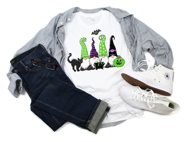May include: White t-shirt featuring a Halloween gnome design with green, purple, and black hats, a black cat, and a green pumpkin. The shirt is paired with blue jeans, a gray jacket, and white sneakers.