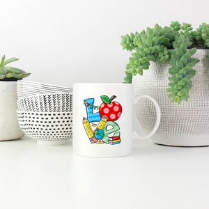 May include: White ceramic mug with a colorful illustration of the word "LOVE" made up of school supplies like pencils, a globe, and a red apple.