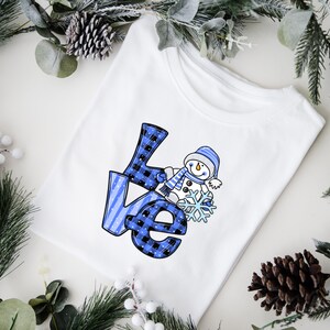 Pu&ograve; includere: T-shirt bianca con la scritta "Love" a quadri blu e neri e un pupazzo di neve che tiene un fiocco di neve. Il pupazzo di neve indossa un cappello e una sciarpa blu. La maglietta &egrave; circondata da verde invernale.