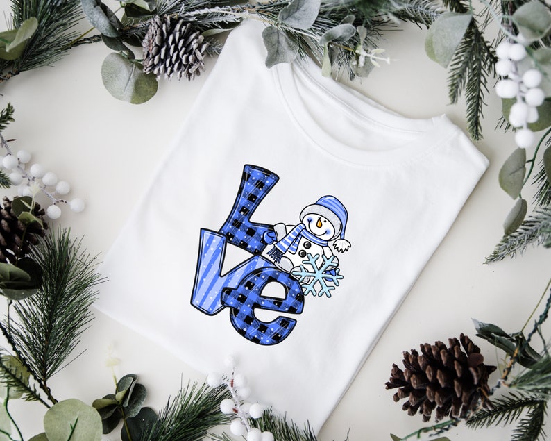 Pu&ograve; includere: T-shirt bianca con la scritta "Love" a quadri blu e neri e un pupazzo di neve che tiene un fiocco di neve. Il pupazzo di neve indossa un cappello e una sciarpa blu. La maglietta &egrave; circondata da verde invernale.