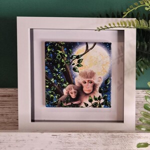 May include: A framed painting depicting two monkeys perched in a tree beneath a full moon. The monkeys are brown and white with black eyes. The background is a dark blue with white stars and green foliage.