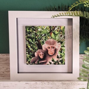 May include: Framed painting of two monkeys in a jungle setting. The mother monkey is brown with a white face and the baby monkey is brown with a white face. The background is a green and brown jungle scene.