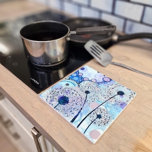 May include: A blue and white ceramic trivet with a dandelion design. The trivet is on a wooden worktop next to a black electric hob.