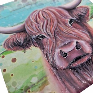 Op de afbeelding: Close-up van een schilderij van een Schotse Hooglandkoe. De koe heeft lang, roodbruin haar, grote donkere ogen en gebogen hoorns. De achtergrond heeft een mix van groene, blauwe en bruine kleuren. Het kunstwerk staat op een vierkante ondergrond.