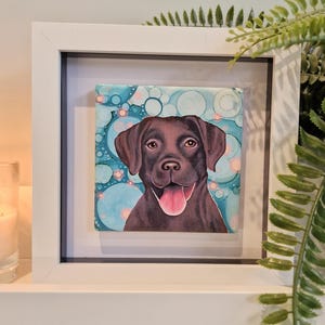 May include: Framed painting of a chocolate brown Labrador Retriever with a pink tongue sticking out. The dog is looking directly at the viewer. The background is a blue and teal abstract design with white circles.