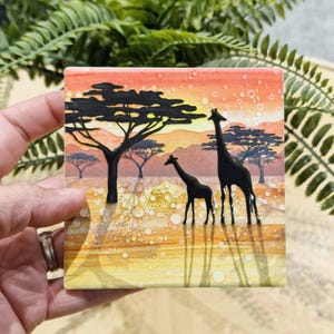 May include: A square tile featuring a sunset scene with two giraffes and acacia trees silhouetted against a yellow, orange, and red sky. The tile has a textured surface with small white dots.