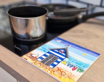 May include: A square trivet with a beach hut design, featuring blue and white stripes, a seagull, and the sea. The trivet is on a wooden surface near a hob with a saucepan and frying pan.