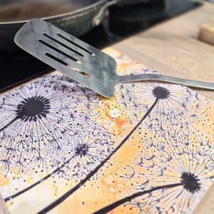 May include: A silver spatula with a black handle sits on a yellow and white trivet with a dandelion design.