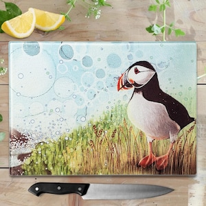 May include: A rectangular glass chopping board with a colourful illustration of a puffin bird in a grassy field. The bird has black, white, and orange markings. A black-handled knife rests below the board. Two lemon slices are visible.
