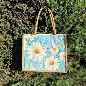 May include: A reusable tote bag made of natural jute fabric with a design of white daisies on a blue background with white circles.