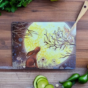 May include: A glass chopping board featuring a charming illustration of a brown rabbit gazing up at a full moon. A white owl sits on a branch bathed in the moon's light. The background is a dark blue with white dandelion-like flowers.