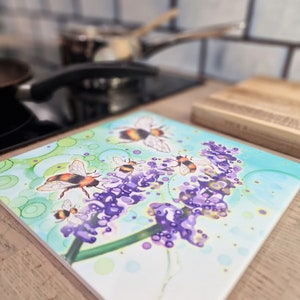 May include: A square ceramic trivet featuring a watercolour design of purple lavender flowers and bumblebees. The trivet has a light blue background with green leaves and white circles.