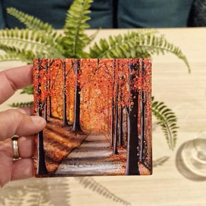 May include: A painting depicting a forest path with trees lining either side. The trees are painted in shades of orange, red, and brown. The path is painted in shades of grey and white. The background is painted in shades of orange and yellow.