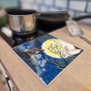 May include: A blue and yellow ceramic tile featuring a white owl and a brown rabbit in front of a full moon. The tile has a dark blue background with white and yellow accents.