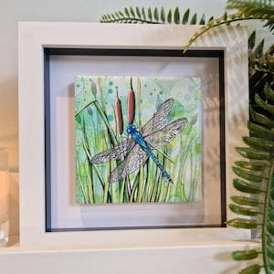 May include: A framed watercolour painting of a blue dragonfly with black and white wings, perched on green reeds with brown cattails. The background is a light blue and green watercolour wash with white dots.