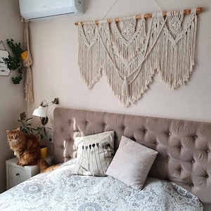 以下が含まれることがあります： ベージュのタフテッドヘッドボードに、白とグレーのパターンの掛け布団をかけたベッドに2つの枕が置かれています。ヘッドボードの上の壁には、木製の棒が付いたマクラメの壁掛けが飾られています。壁は薄いピンク色です。