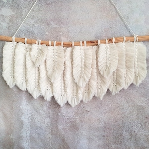 May include: A white macrame wall hanging with a wooden dowel rod and a fringe of white yarn. The wall hanging is shaped like a series of feathers.