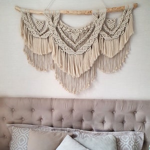 May include: A beige macrame wall hanging with a wooden branch at the top. The wall hanging is made of knotted cords and has a fringe at the bottom. The wall hanging is hanging above a beige headboard with a tufted design.