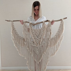 May include: A large, beige macrame wall hanging with a geometric pattern and fringe. The hanging is attached to a natural wood branch.