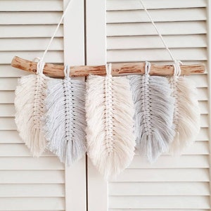 May include: A wall hanging made of macrame feathers in shades of white and gray. The feathers are attached to a wooden branch.