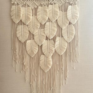 May include: Cream-colored macrame wall hanging featuring a series of leaf-shaped designs. The leaves are arranged in rows and suspended from a wooden dowel. The piece has a textured, handmade appearance, with long, dangling fringe.